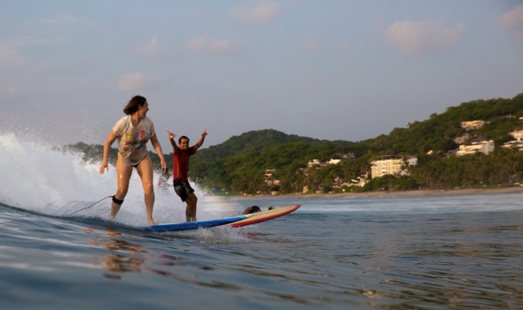 A student riding a wave while Carlos cheers behind her
